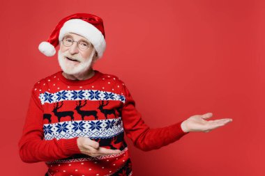 Süveter ve Noel Baba şapkalı, elleri kırmızı arka planda gülümseyen yaşlı adam.