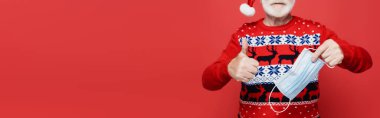 Kırmızı pankartta izole edilmiş, bulanık arka planda tıbbi maske tutan, Noel Baba şapkalı adam görüntüsü. 