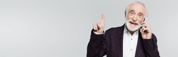 Улыбающийся пожилой человек указывает пальцем, разговаривая на смартфоне, изолированном на сером, баннер
