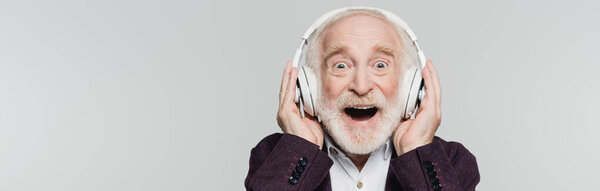 Excited senior man listening music in wireless headphones isolated on grey, banner 
