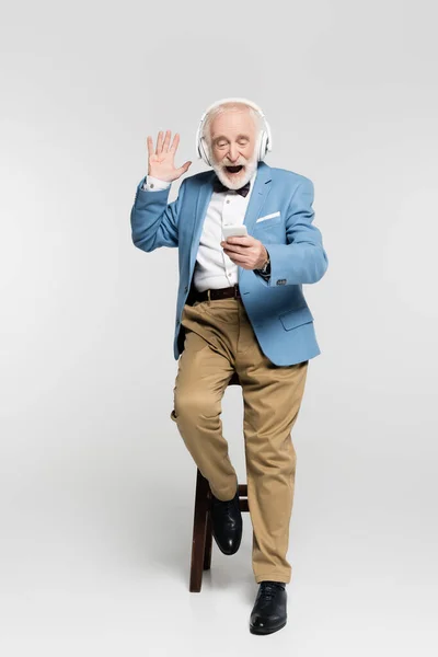 興奮したシニア男でヘッドフォン手で手を振ってスマートフォン上の椅子にグレーの背景 — ストック写真