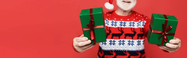 Cropped view of gifts in hands of senior man in santa hat on blurred background isolated on red, banner