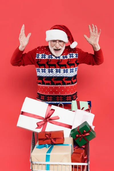 Excited senior man in santa hat looking at gifts in shopping cart isolated on red