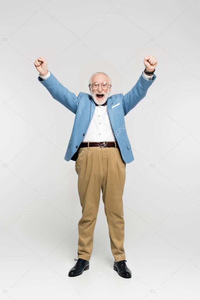 Excited senior man in blue jacket and eyeglasses showing yes gesture on grey background 