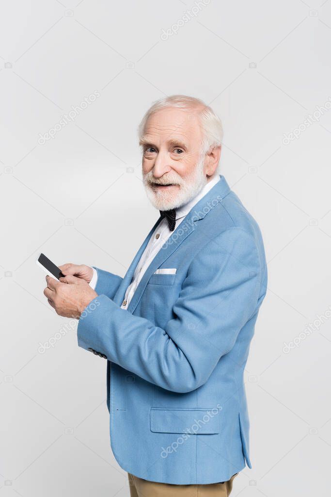 Senior man in shirt and jacket holding smartphone and smiling at camera isolated on grey 