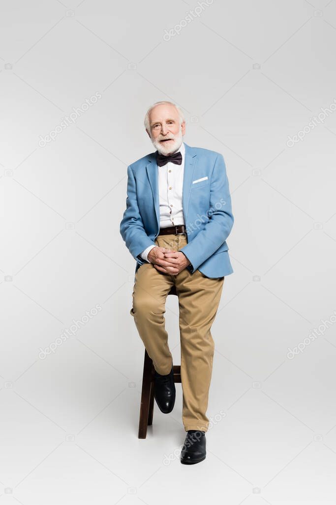 Senior man in bow tie and blue jacket sitting on chair on grey background 