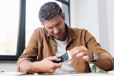 Irritated businessman with smartphone removing damaged touchscreen with blurred window on background clipart