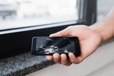 Cropped view of businessman holding smashed smartphone on blurred background clipart