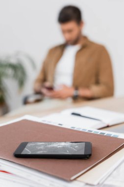 Arka planda bulanık iş adamıyla masadaki kağıt yığınında kırılmış akıllı telefon.
