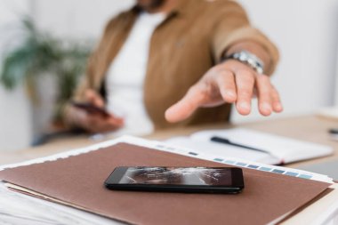İşadamı eli, iş yerindeki bulanık arka plandaki kağıt yığınının üzerinde ezilmiş bir akıllı telefonun yanında görüntülenmiş.