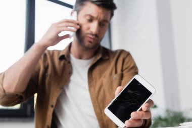 Arkaplanda bulanık bir pencereyle cep telefonuyla konuşan işadamının elinde kırılmış bir akıllı telefon.
