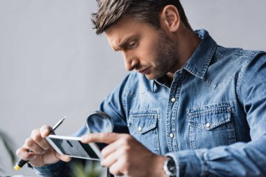 Odaklanmış tamirci, parçalanmış cep telefonundaki büyüteçle ön plandaki parçalanmış görüntüyü inceliyor.