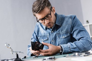 İş yerinde bulanık ön planda büyüteçle otururken bozuk akıllı telefonu parçalayan odaklı tamirci