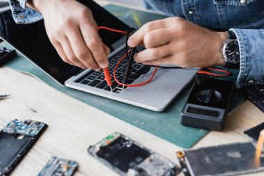 Cropped view of repairman holding sensors of multimeter on broken keyboard of laptop with blurred disassembled phones on foreground clipart