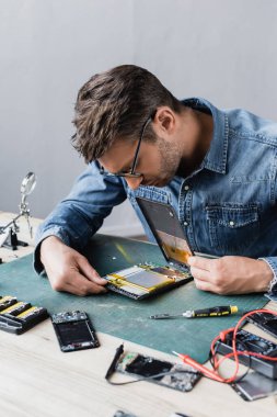 Repairman in eyeglasses looking at disassembled broken digital tablet near screwdriver and multimeter at workplace clipart