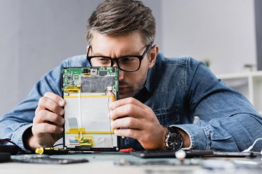 Focused repairman squinting, while looking at part of broken digital tablet with blurred workplace on foreground clipart