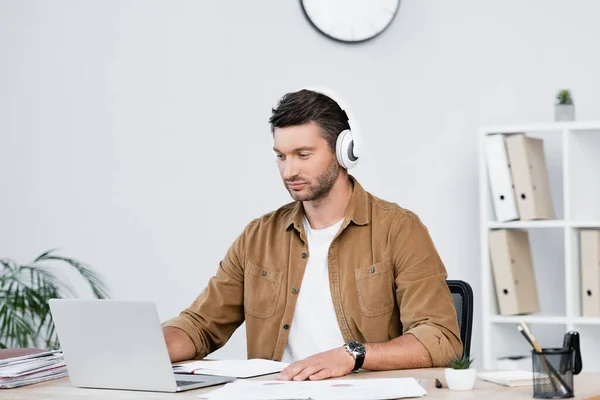 オフィスの職場に座っている間ノートパソコンを見ているヘッドフォンのビジネスマン — ストック写真