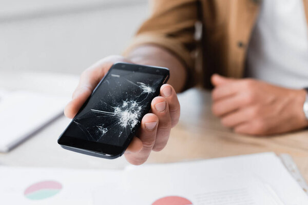 Cropped view of man holding smashed smartphone with blurred workplace on background