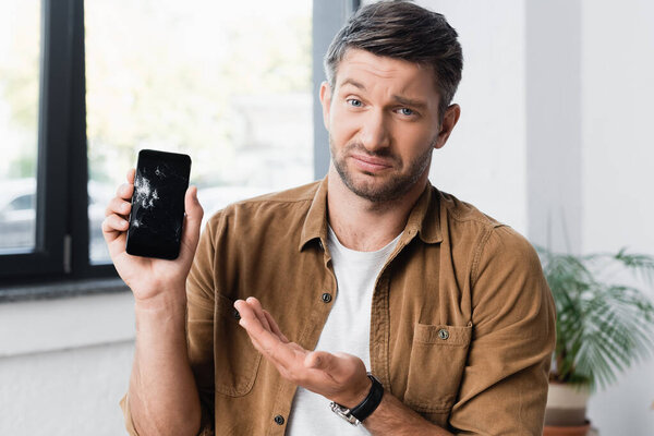 Skeptical businessman looking at camera while pointing with hand at smashed smartphone on blurred background