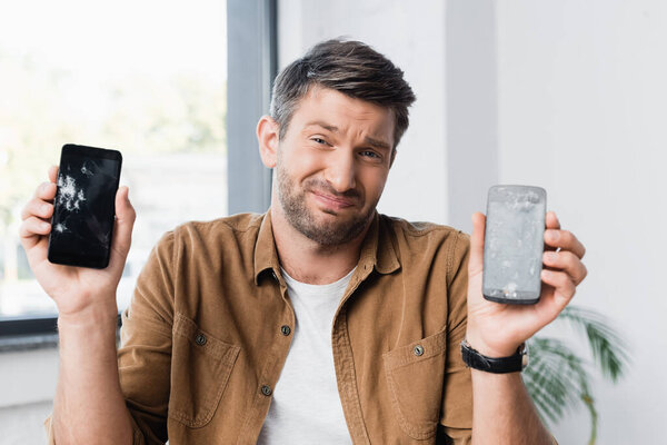 Confused businessman with shrug gesture looking at camera while holding smashed smartphones on blurred background