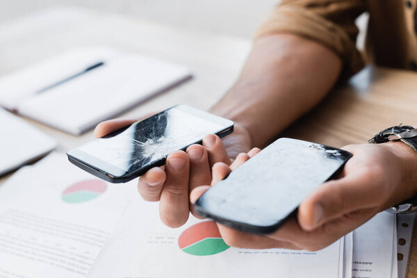 Cropped view of businessman holding damaged smartphones near table on blurred background