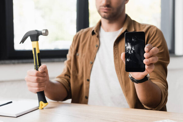 Cropped view of businessman with hammer showing damaged smartphone while sitting at workplace on blurred background