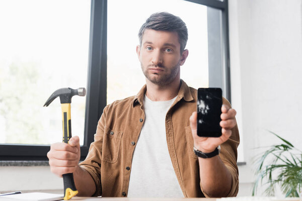 Sad businessman with hammer looking at camera while showing damaged smartphone with blurred window on background