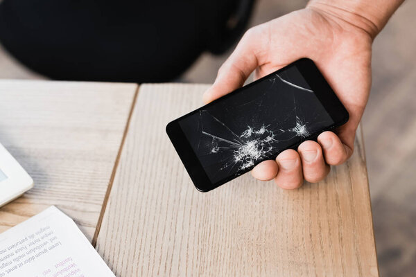 Обрезанный вид человека с разбитым смартфоном рядом со столом на размытом фоне