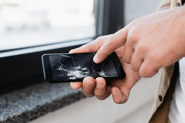 Cropped view of businessman touching smashed touchscreen of smartphone on blurred background