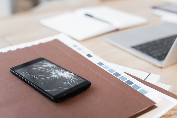 Close up view of smashed smartphone on pile of paperwork with blurred workplace on background