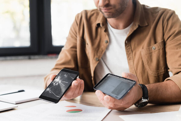 Cropped view of businessman holding disassembled damaged smartphone at workplace on blurred background