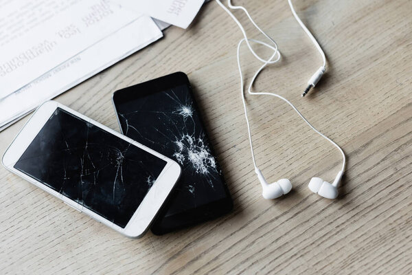 Top view of earphones and smashed smartphones near documents on wooden background, banner