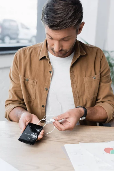 Businessman with earphones, holding smashed smartphone while sitting at workplace on blurred background