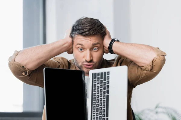 Mad businessman with hands near head looking at damaged laptop on blurred background