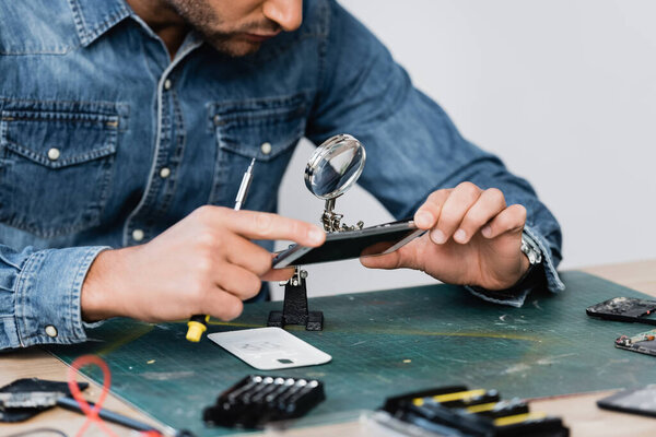 Cropped view of repairman looking through magnifier at disassembled part of cellphone at workplace on blurred foreground