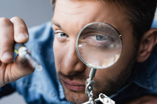 Close up view of focused repairman looking through magnifier at screwdriver on blurred foreground