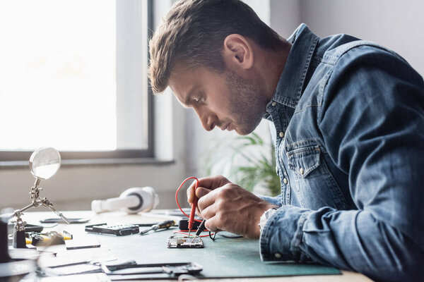 Side view of focused repairman holding sensors of multimeter on disassembled part of mobile phone on blurred background