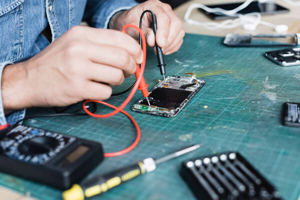 Cropped view of repairman holding multimeter sensors on disassembled part of mobile phone on blurred foreground