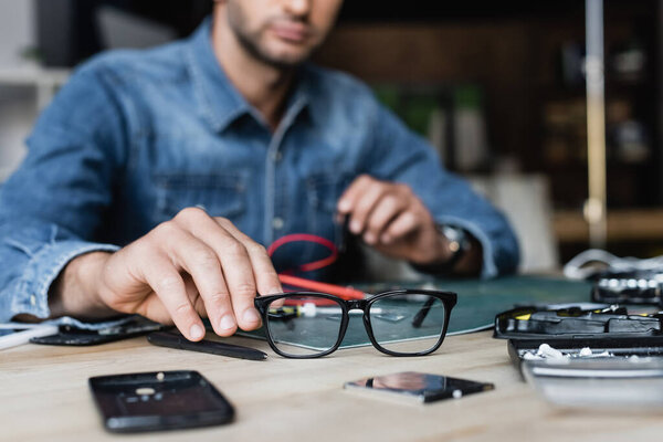 Cropped view of repairman taking eyeglasses from table with disassembled parts of mobile phones on blurred background