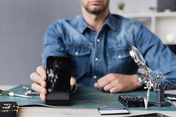 Cropped view of repairman showing smashed smartphone while sitting at table with magnifier on blurred background