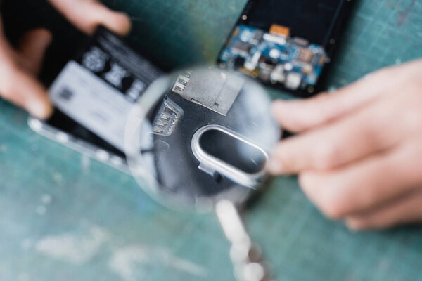 Cropped view of repairman holding magnifier over broken parts of mobile phones on blurred foreground