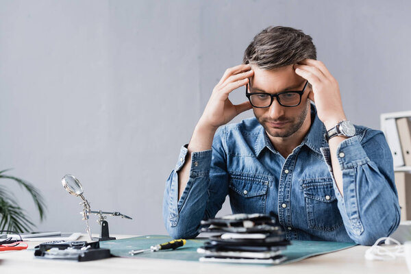 Tired repairman with hands near head looking at pile of broken mobile phones on blurred foreground