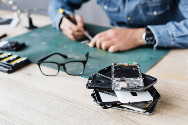 Close up view of pile with broken mobile phones near eyeglasses on workplace with blurred repairman working on background