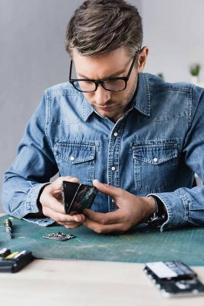 Focused repairman disassembling broken smartphone on workplace on blurred foreground