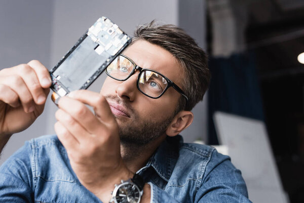 Focused repairman in eyeglasses looking at disassembled component of mobile phone on blurred background