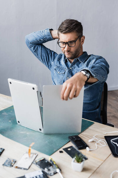 Thoughtful repairman with hand behind head looking at broken laptop at workplace on blurred foreground