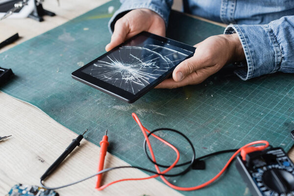 Cropped view of repairman holding smashed digital tablet near multimeter at workplace 