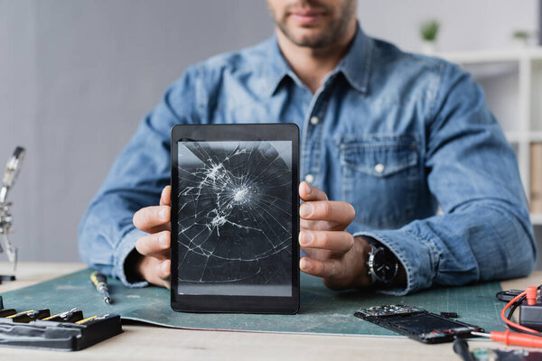 Cropped view of repairman showing smashed digital tablet, while sitting at workplace 