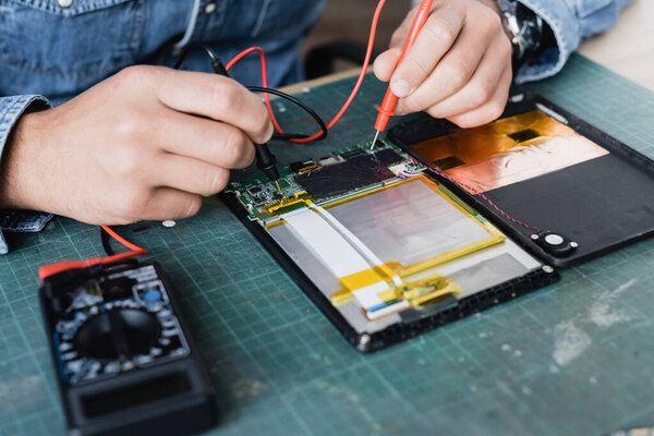 Close up view of repairman hands holding sensors of multimeter on part of broken digital tablet at workplace