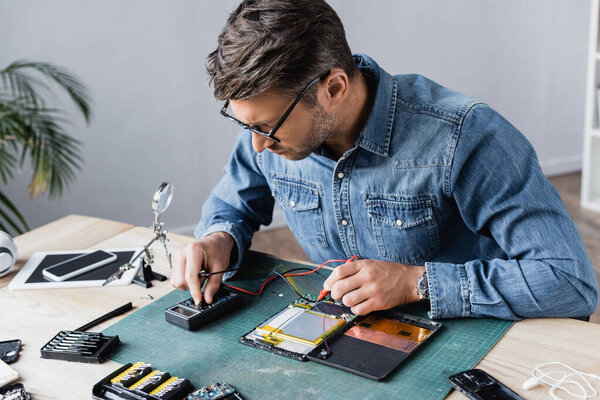 Repairman regulating multimeter while holding sensors near disassembled part of broken digital tablet at workplace